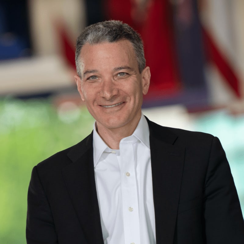 A smiling man in a black suit and white shirt is in the foreground, with a blurred background of red, white, and blue elements.