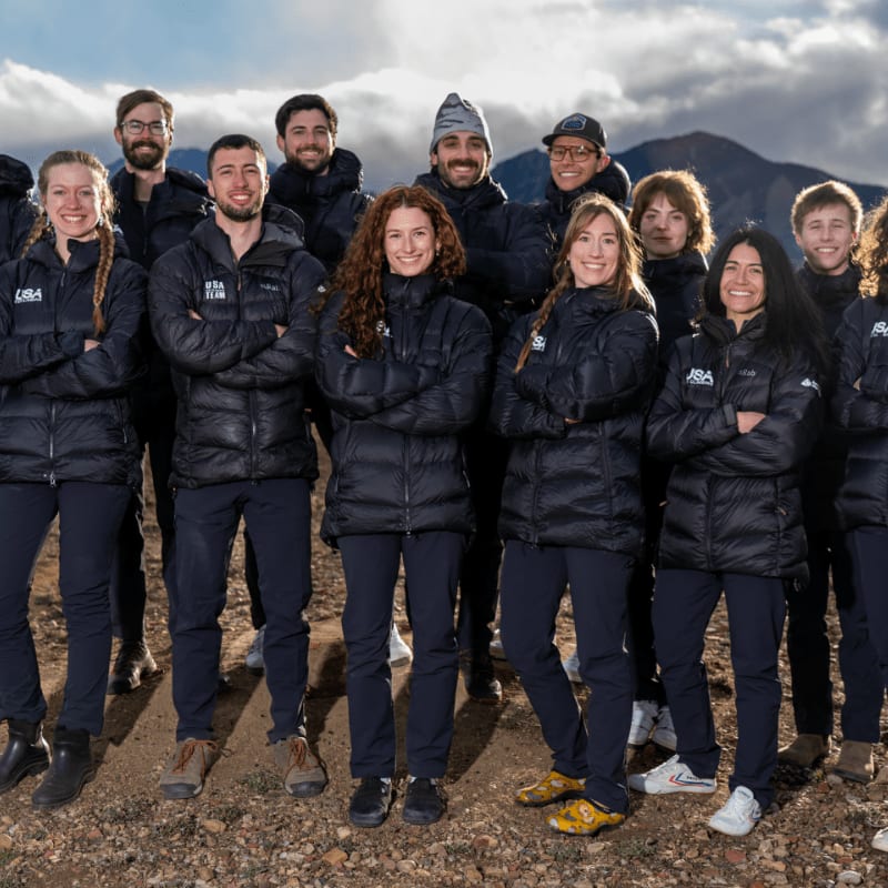 A group of people in puffy jackets stand in the foreground with mountains in the background.