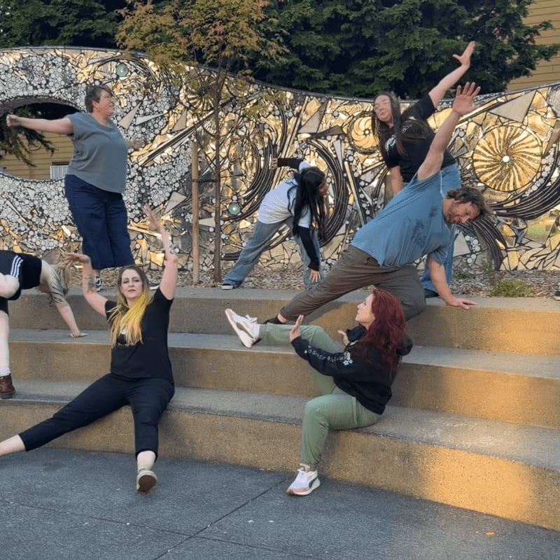 A group of people in various yoga poses are on concrete steps in front of a mosaic wall.