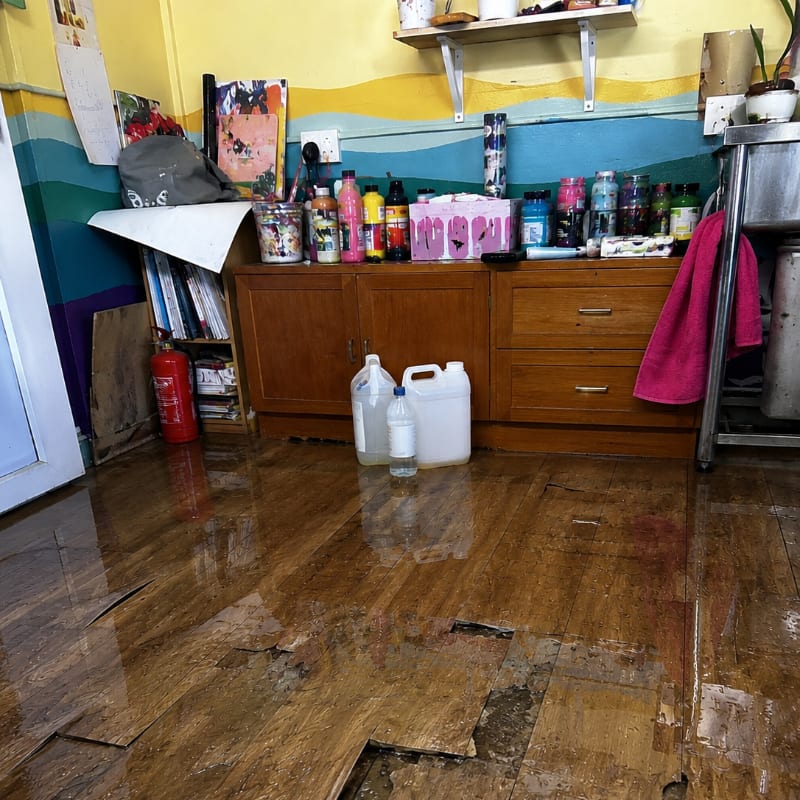 Water-damaged flooring with peeling laminate is visible in the foreground, while the background features a cluttered counter with bottles and a sink.