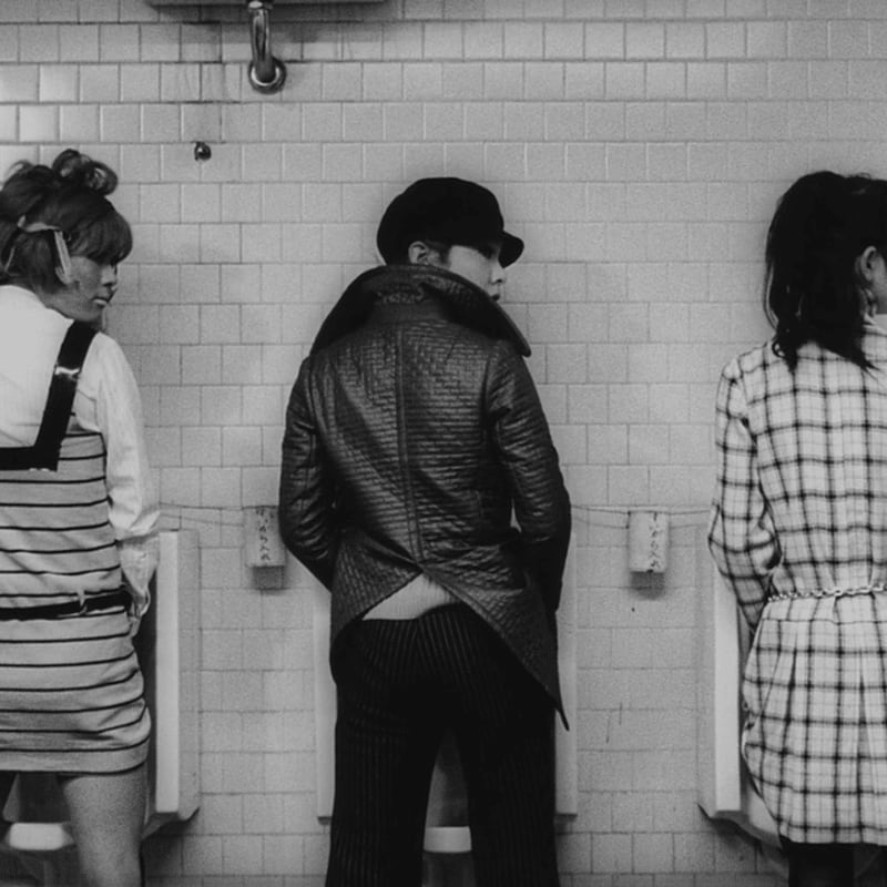 Three people stand at urinals in a tiled restroom, with the central figure wearing a hat and quilted jacket.