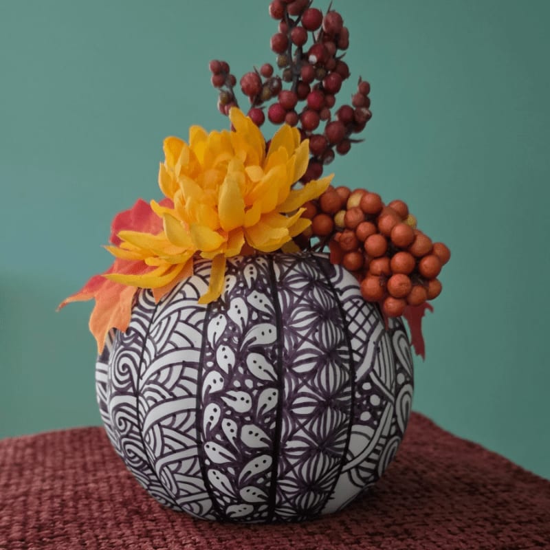 A black and white patterned pumpkin vase filled with autumn flowers and berries sits on a textured red cloth against a teal background.