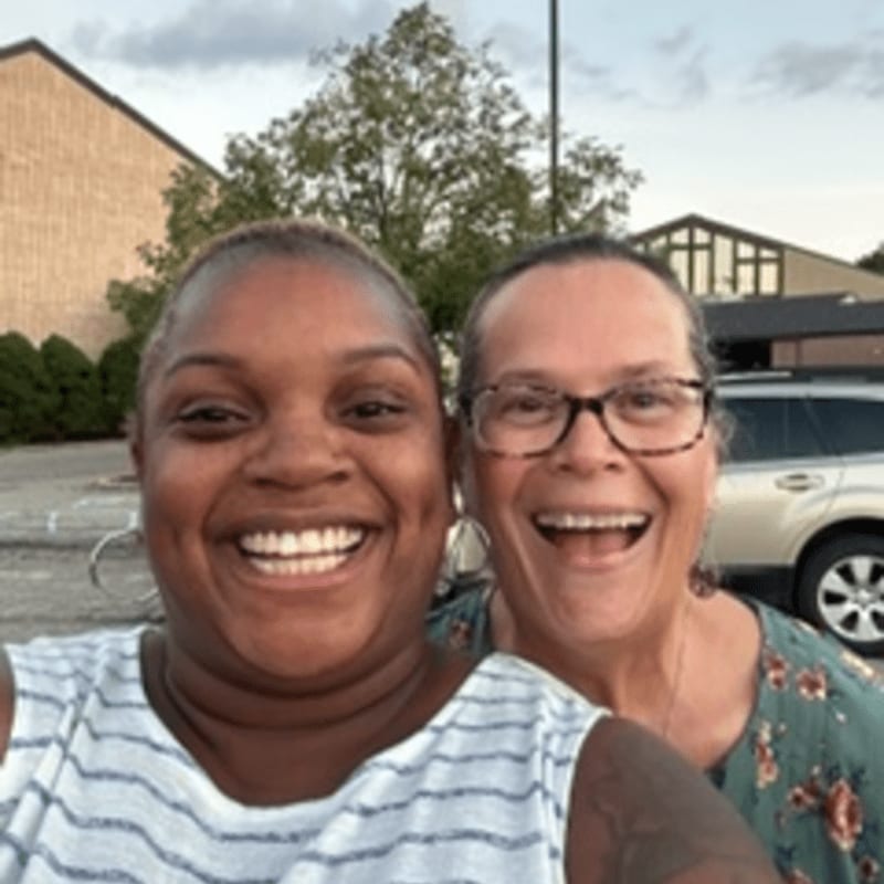 Two smiling women are in the foreground, with a building and a car visible in the background.