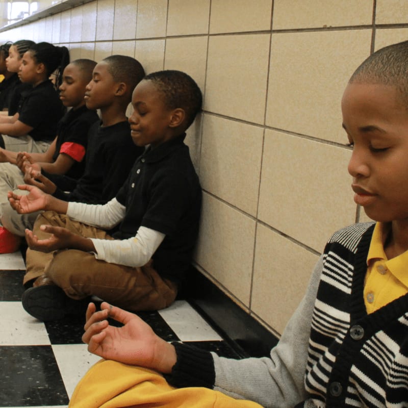 A group of young students in uniform sit in meditation with their eyes closed, against a tiled wall in a hallway.