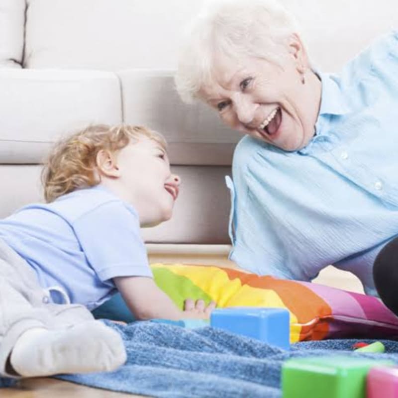 A smiling grandmother and a young child with his tongue out play with colorful toys on the floor in front of a couch.