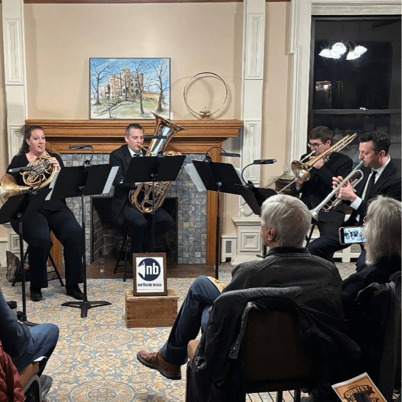 A brass quintet performs in front of a fireplace with a painting above it, while an audience watches from chairs in the foreground.