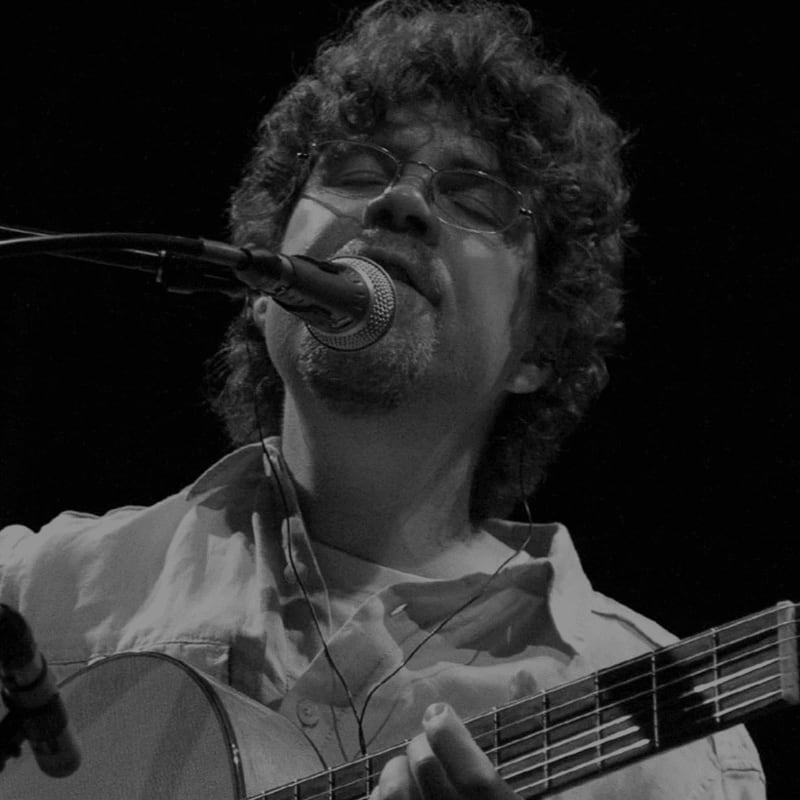 A man with curly hair and glasses sings into a microphone while playing a guitar, with a dark background.