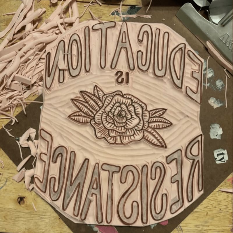 A linocut block with the words "EDUCATION IS RESISTANCE" carved around a rose is surrounded by carved linoleum scraps on a wooden surface.