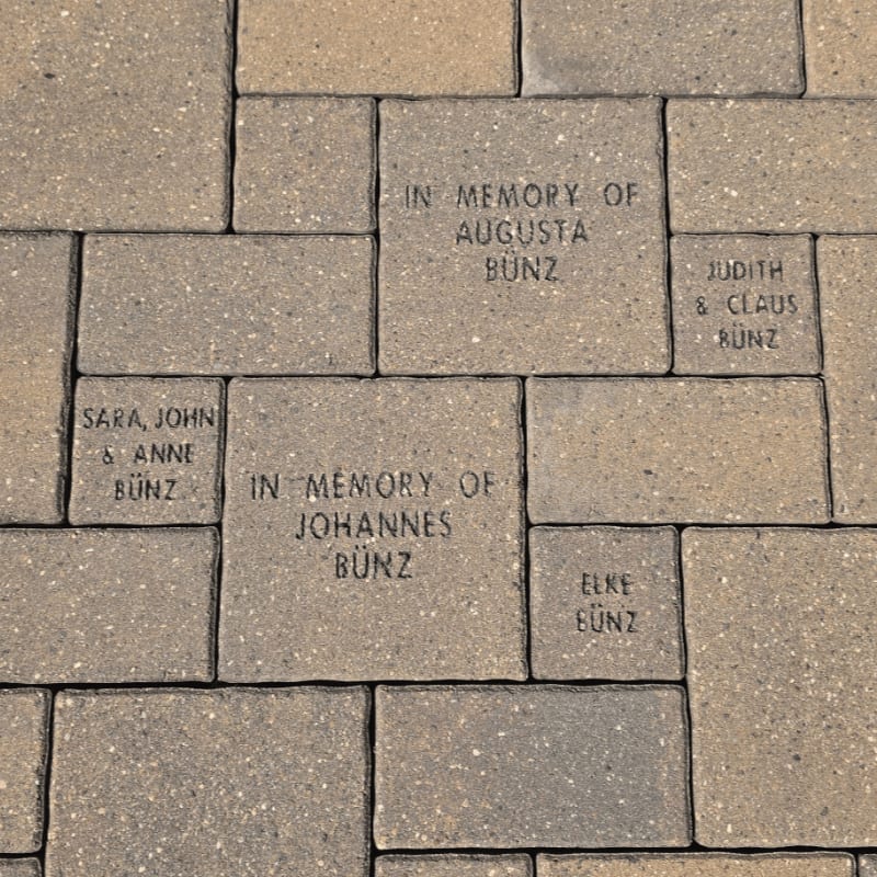 Engraved memorial bricks form a textured ground in the foreground, with names like Augusta, Johannes, and Elke Bunz visible, set against a backdrop of more of the same paving stones.
