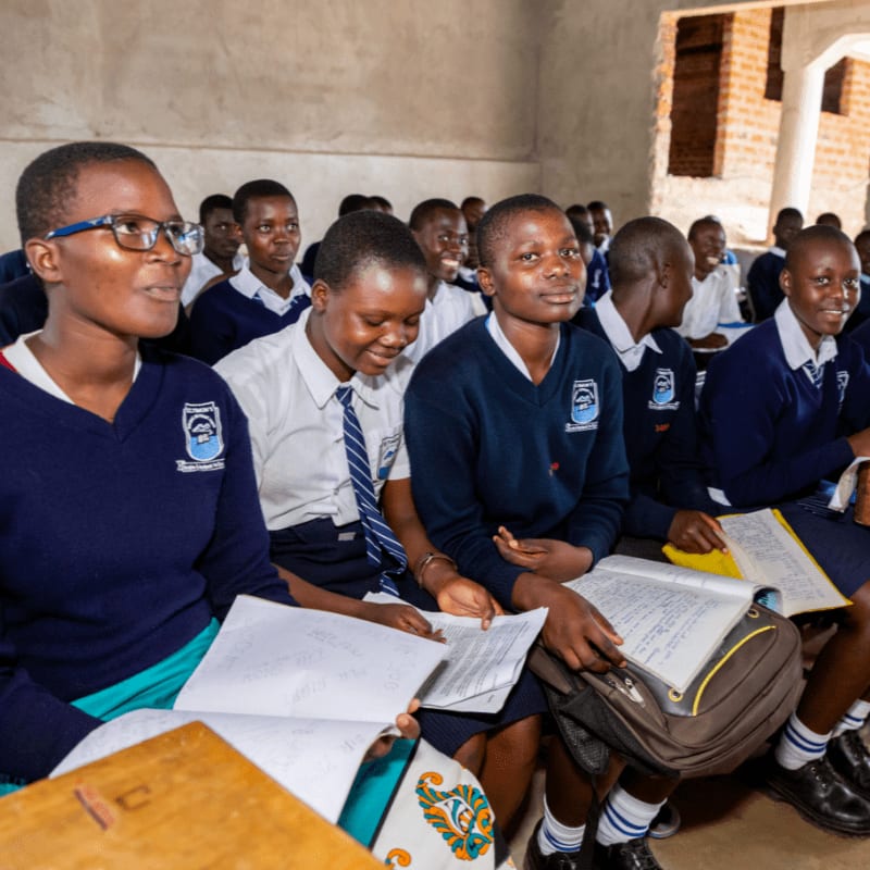 Students in school uniforms sit in a classroom, with some reading books and others looking forward, while the background shows more students and the classroom's brick walls and pillars.