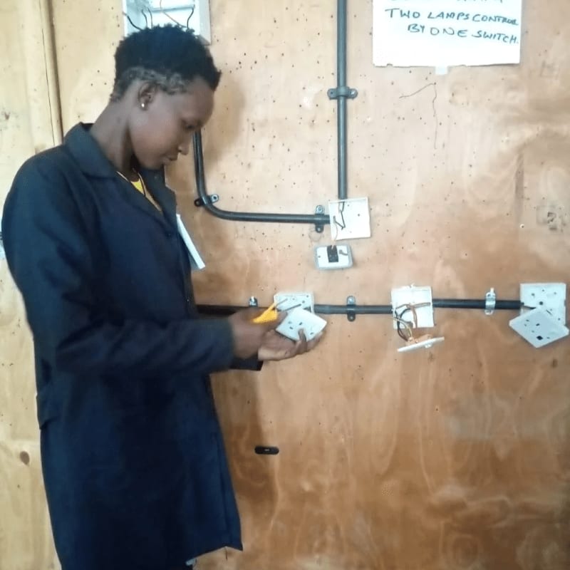 A young person in a dark blue uniform works on electrical outlets and wiring on a wooden wall, with a sign in the background indicating "TWO LAMPS CONTROLLED BY ONE SWITCH."