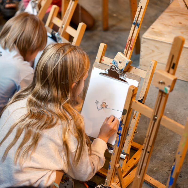 A young girl with long blonde hair draws on an easel in the foreground, while another child is visible in the background.