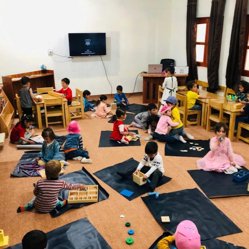 Children are engaged in various activities on the floor in the foreground, while others are seated at tables in the background of a classroom.
