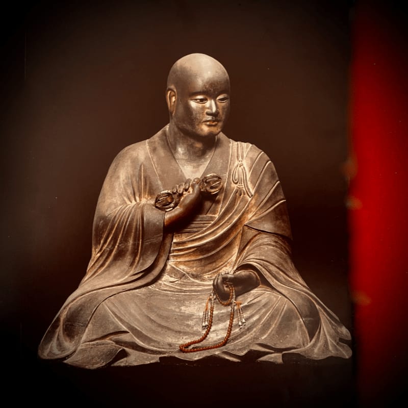 A multi-armed statue with a fierce expression sits in meditation on a lotus throne against a dark background.