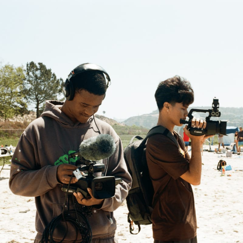 Two young men with video cameras, one wearing headphones, are filming on a sandy beach with hills and trees in the background.