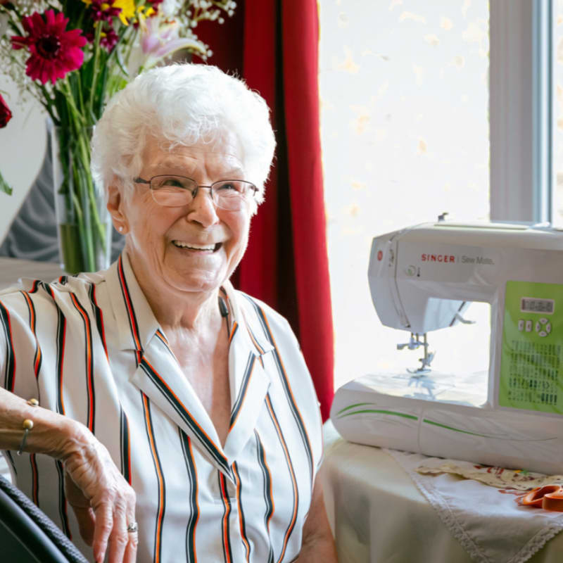 A smiling elderly woman with glasses sits in the foreground next to a sewing machine, with a bouquet of flowers and red curtains in the background.
