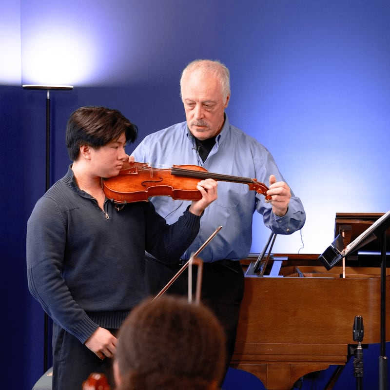 A young man plays the violin while an older man instructs him, with a piano and music stand in the background.
