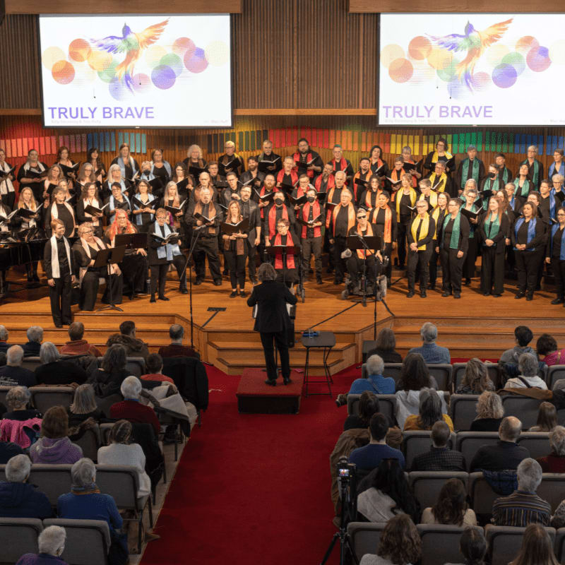 A choir performs on a stage with a conductor in the foreground, while an audience watches from tiered seating in the background.