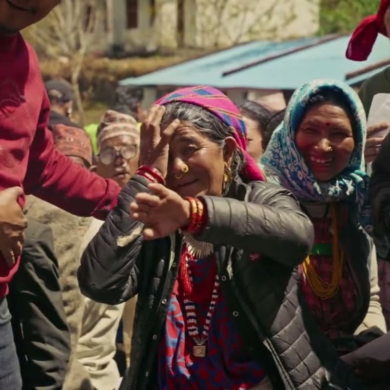 An elderly woman in the foreground with her hand on her forehead is surrounded by smiling people in the background, with buildings visible behind them.
