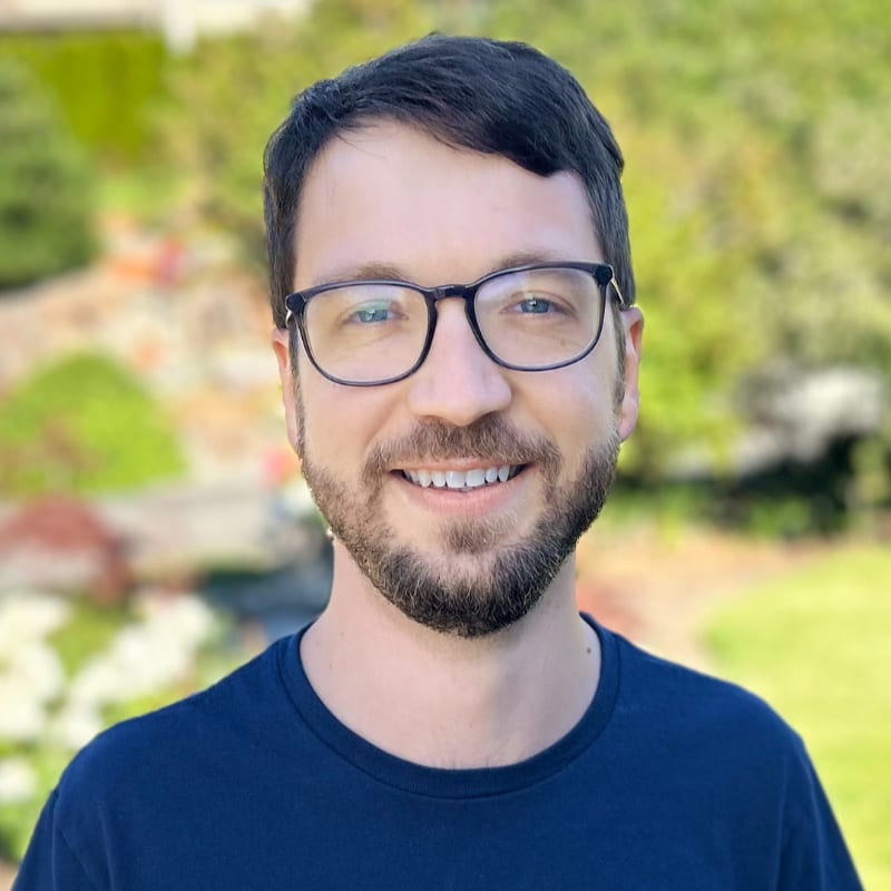 A smiling man with glasses and a beard in a dark blue shirt is in the foreground, with a blurred background of greenery and a stone wall.