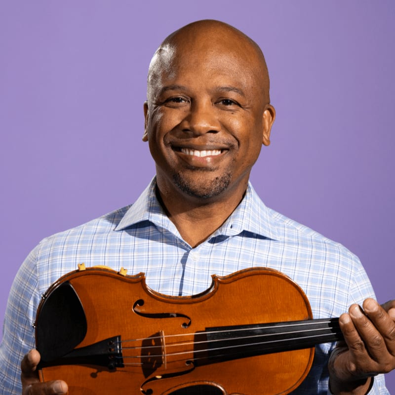 A smiling bald man in a blue checkered shirt holds a violin in front of a solid purple background.