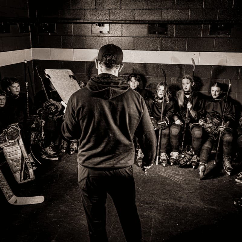 A coach in a hoodie addresses a group of young hockey players sitting on a bench with their gear in a locker room.