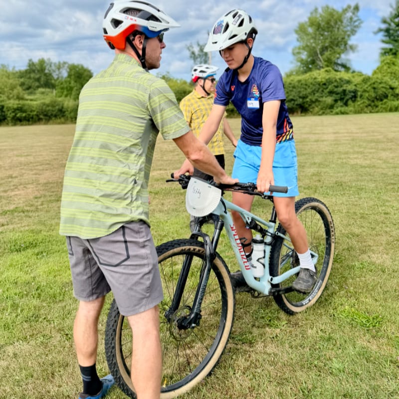 A man in a green shirt and grey shorts helps a young person in a blue shirt and blue shorts learn to ride a bicycle on a grassy field with trees in the background.