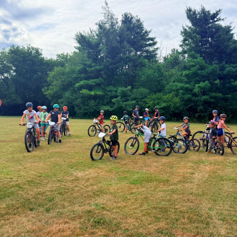 A group of children on bicycles stand on a dry, grassy field with a line of trees in the background under a cloudy sky.