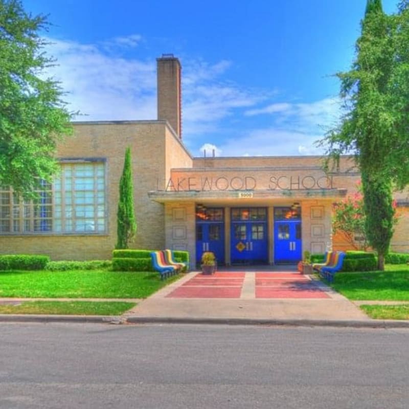 Brightly colored benches and manicured bushes line the sidewalk in front of the Lakewood School, a tan brick building with blue double doors, set against a clear blue sky.