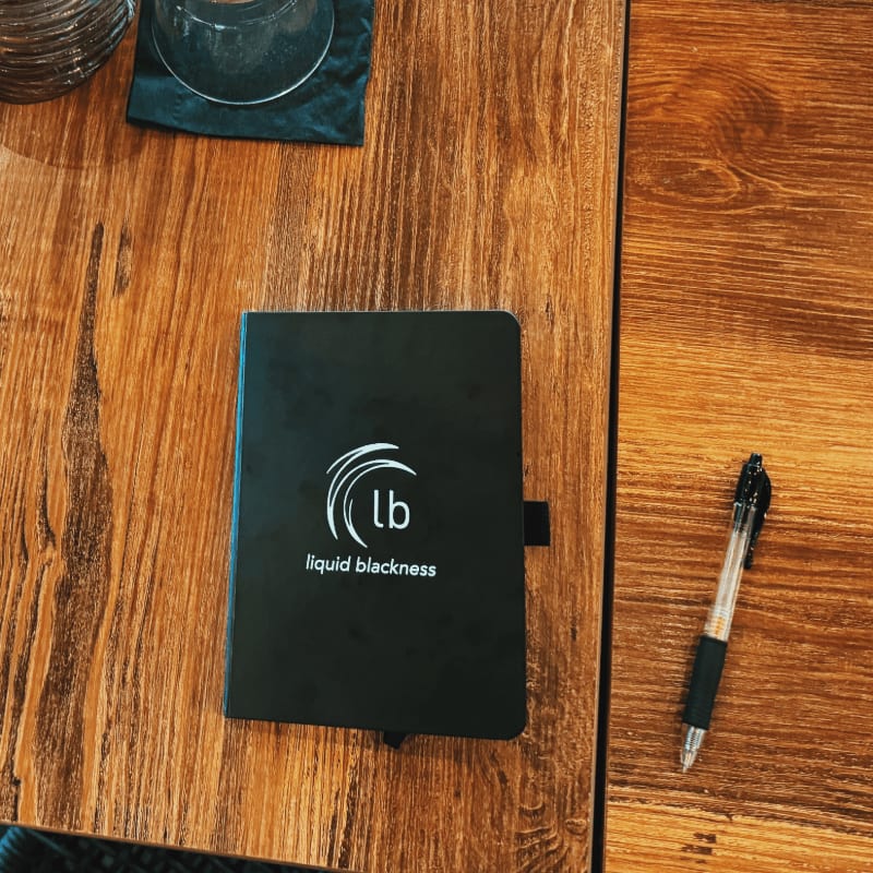 A black notebook with a logo sits on a wooden table next to a pen, with a glass and coaster visible in the upper left corner.