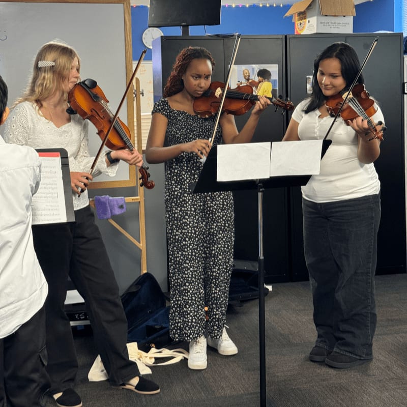 Three young women play violins in a classroom with a whiteboard and cabinets in the background.
