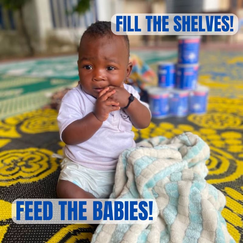A baby sits on a patterned mat in the foreground with a blanket, while cans of food are stacked in the background under a blue banner that reads "FILL THE SHELVES!"