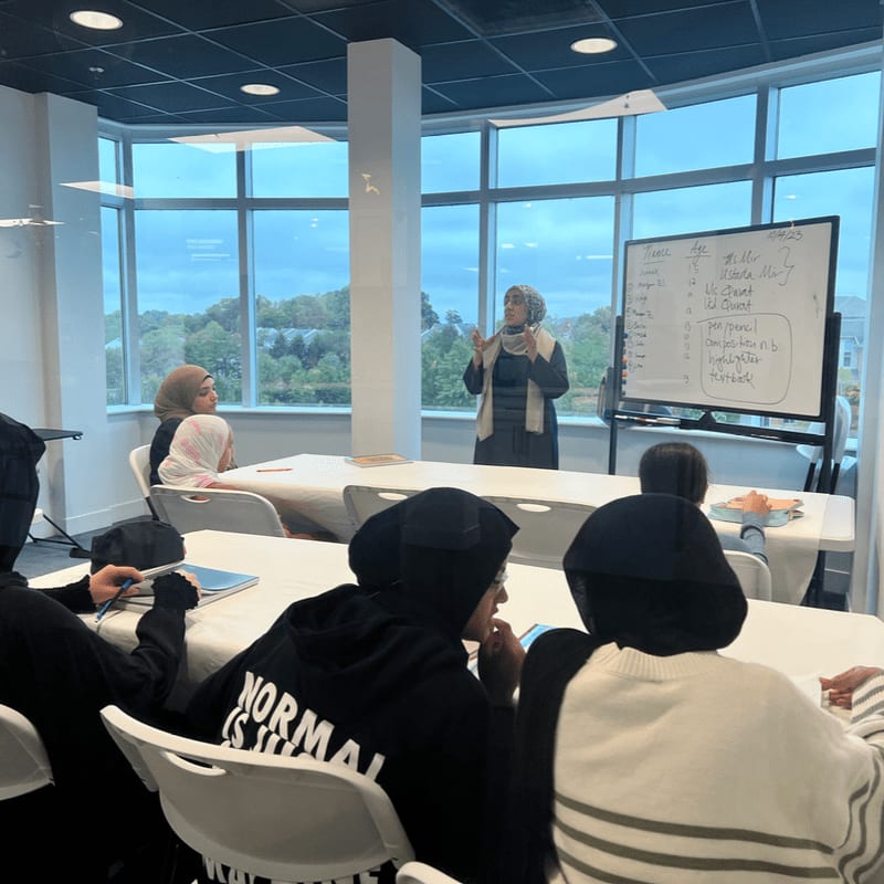 In the foreground, students wearing hijabs are seated at tables, while in the background, a woman stands by a whiteboard in a brightly lit room with large windows overlooking trees.