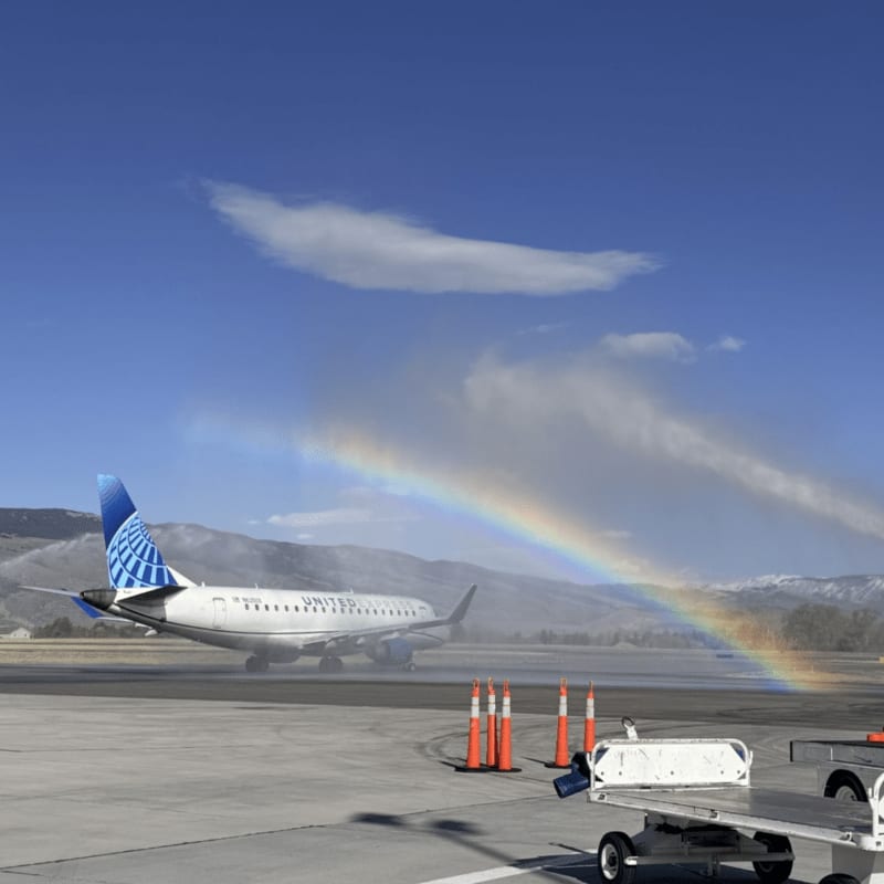 A United Airlines plane is greeted with a rainbow water salute on an airport tarmac, with mountains in the background under a blue sky.