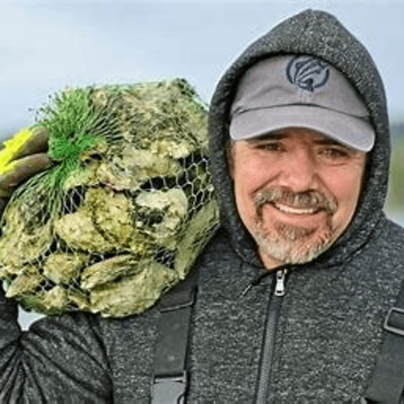 A man in a gray hoodie and baseball cap smiles as he carries a net bag full of oysters over his shoulder, with a blurred outdoor background.