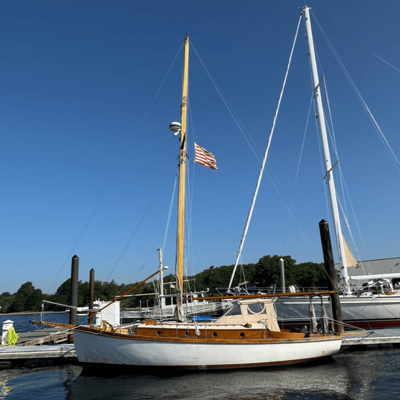 A classic wooden sailboat with a tan cabin and an American flag is docked in the foreground, with a larger white sailboat and a tree-lined shore visible in the background under a clear blue sky.