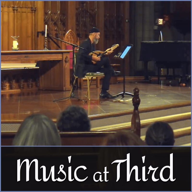 A musician plays a mandolin on a stage in front of an altar and pews, with an audience visible in the foreground.