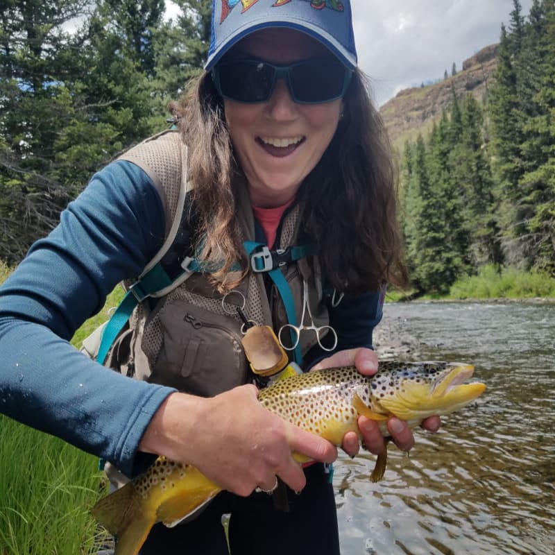 A smiling woman in fishing gear holds a brown trout in the foreground, with a river and pine-covered hills in the background.