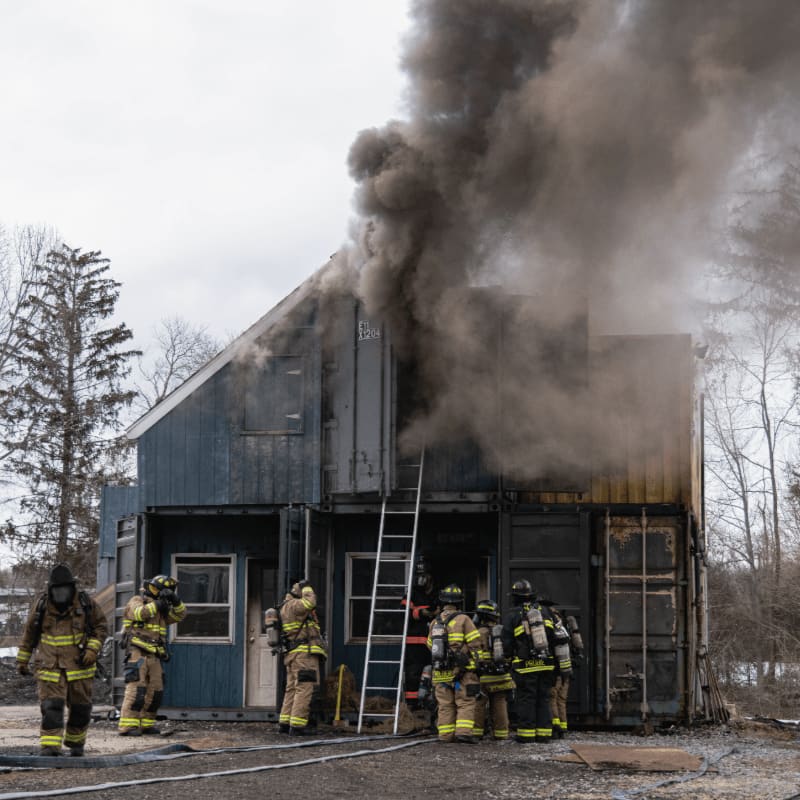 Firefighters in protective gear surround a burning building with thick smoke billowing from its roof, while bare trees and a cloudy sky form the background.