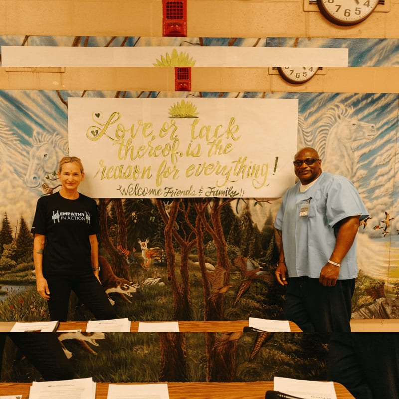 A woman in a black t-shirt and a man in a light blue uniform stand behind a table with papers, in front of a mural depicting a forest scene with animals and a large quote about love.
