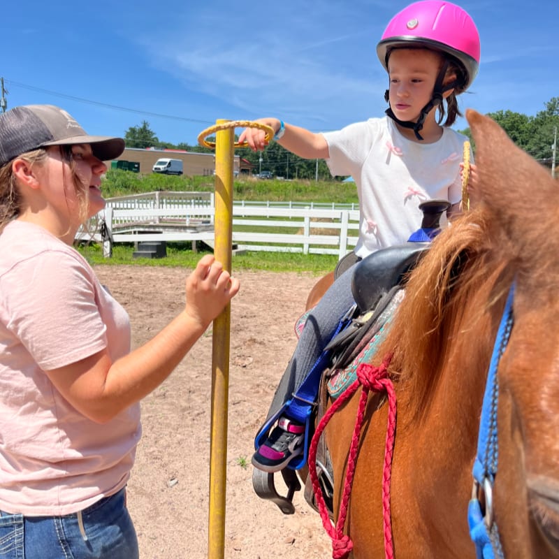 A young girl in a pink helmet rides a horse while a woman in a hat holds a yellow pole, with a fenced area and trees in the background.