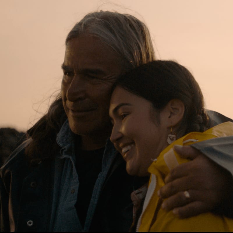 A man with long gray hair embraces a smiling young woman in a yellow jacket, with a soft, hazy sky in the background.