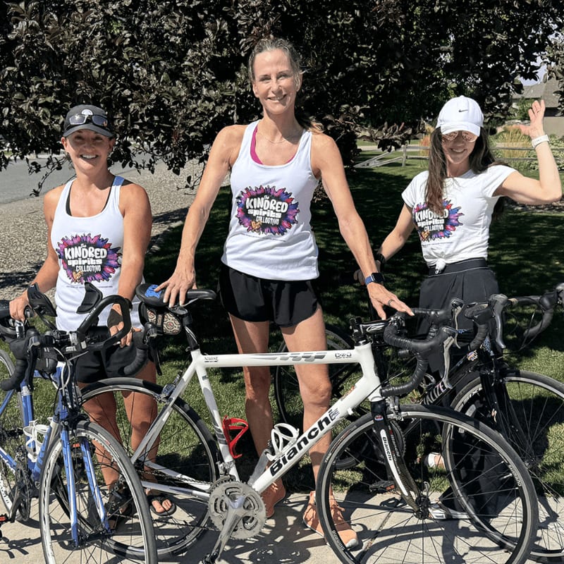 Three women in matching "Kindred Spirits" tank tops pose with their bicycles in front of a tree-lined street.