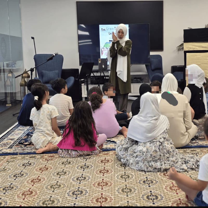 Children sit on a patterned rug in the foreground, listening to a woman in a hijab read from a book on a screen in the background.