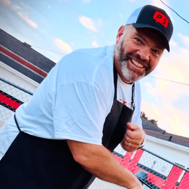 A smiling man in a white t-shirt and black apron, wearing a black and white trucker hat, poses in front of a building with red and white striped accents under a cloudy sky.