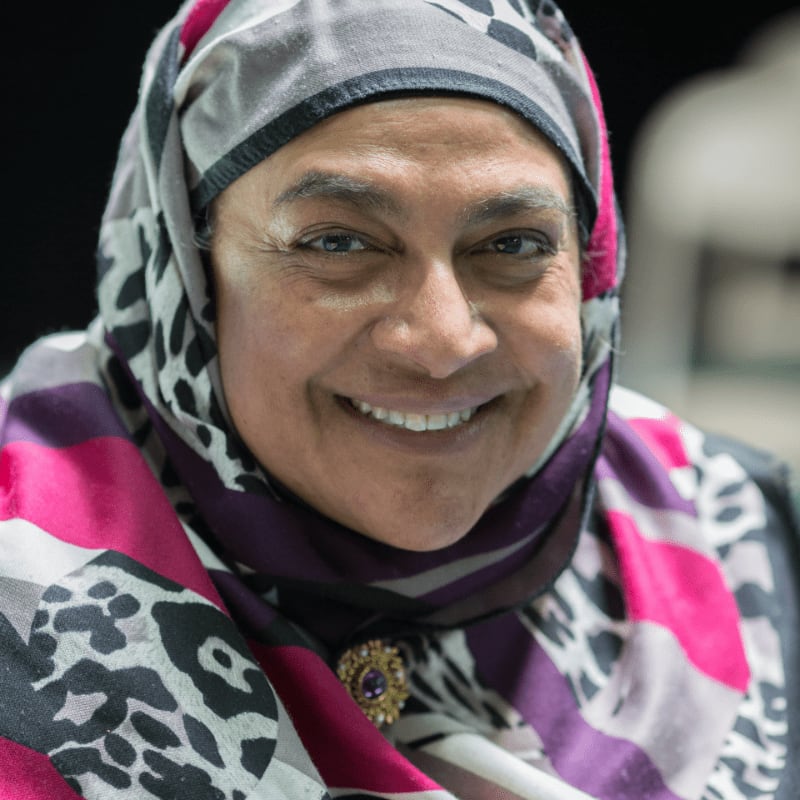 A smiling woman in a colorful hijab is in the foreground, with a blurred, dark background.