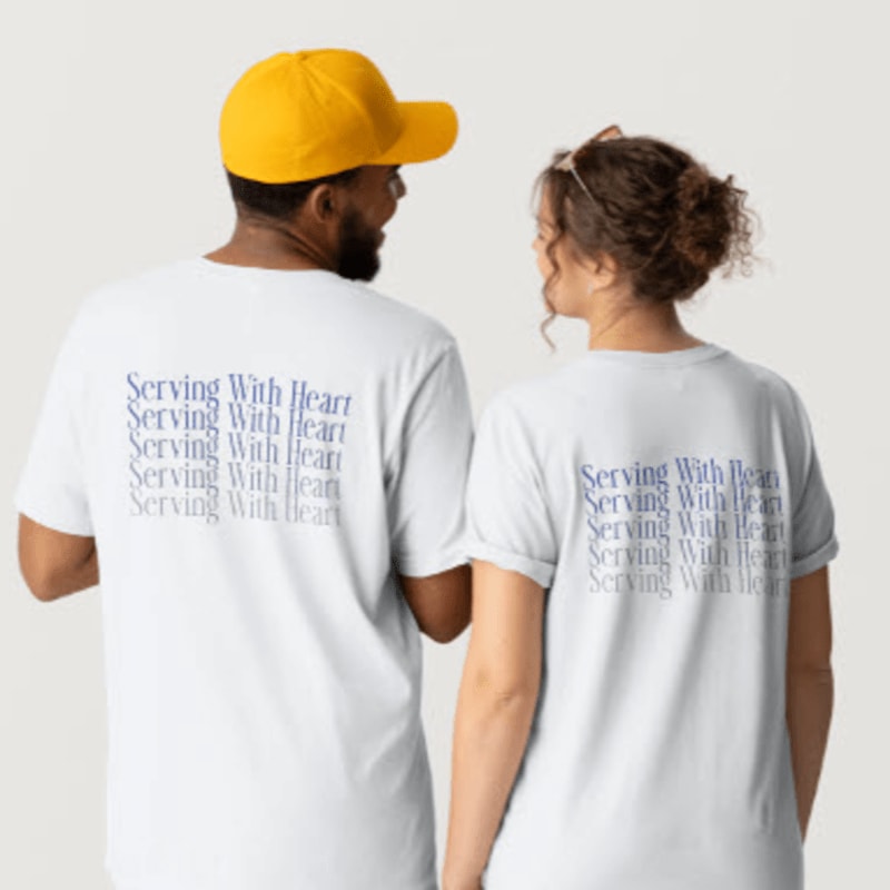 A man wearing a yellow cap and a woman stand side-by-side, both with their backs to the camera, showcasing white t-shirts with the phrase "Serving With Heart" printed repeatedly in blue on the back.