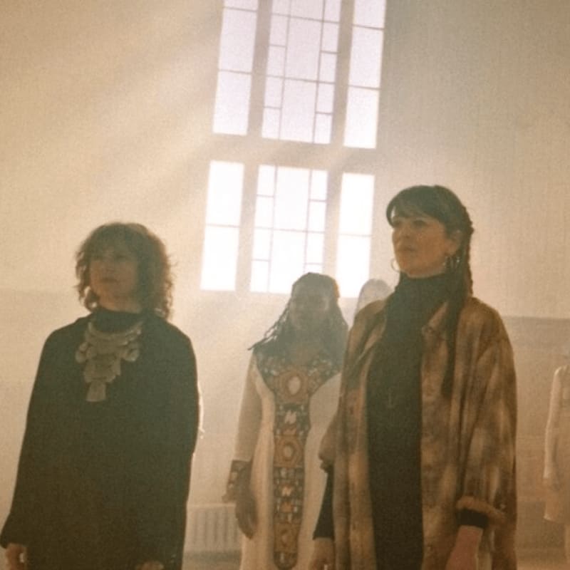 Three women stand in a dimly lit room with a large window in the background.