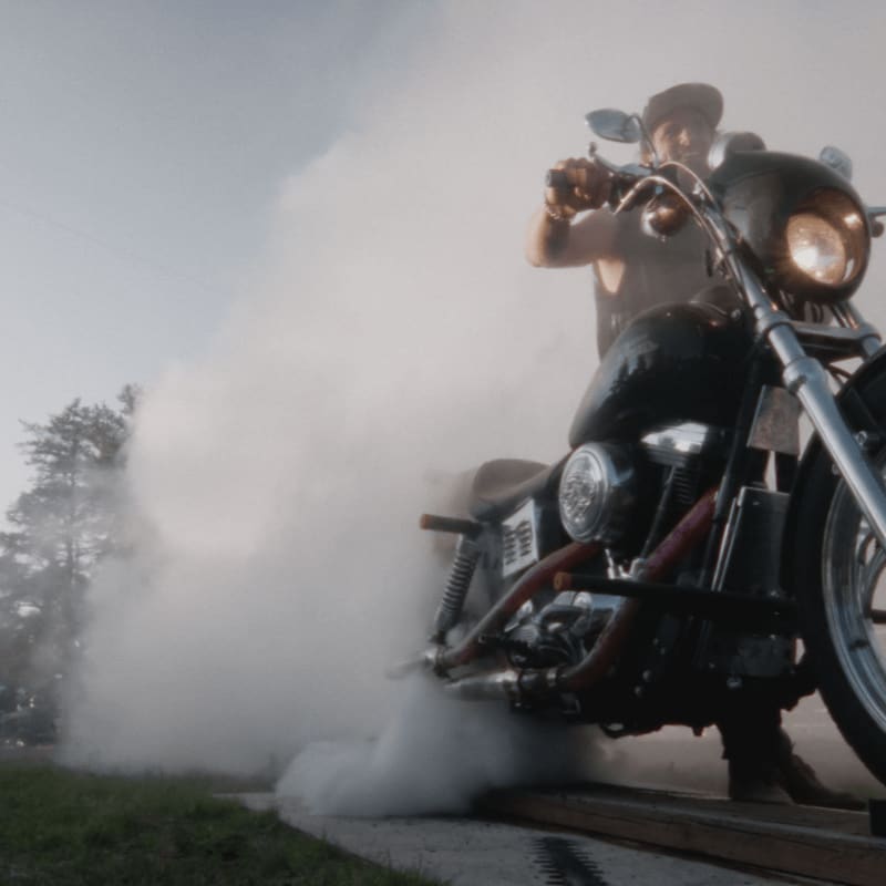 A man on a motorcycle creates a large cloud of smoke in the foreground, with trees and a cloudy sky in the background.