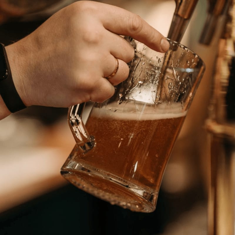 A hand with a ring pours amber beer into a glass mug, with a blurred bar in the background.